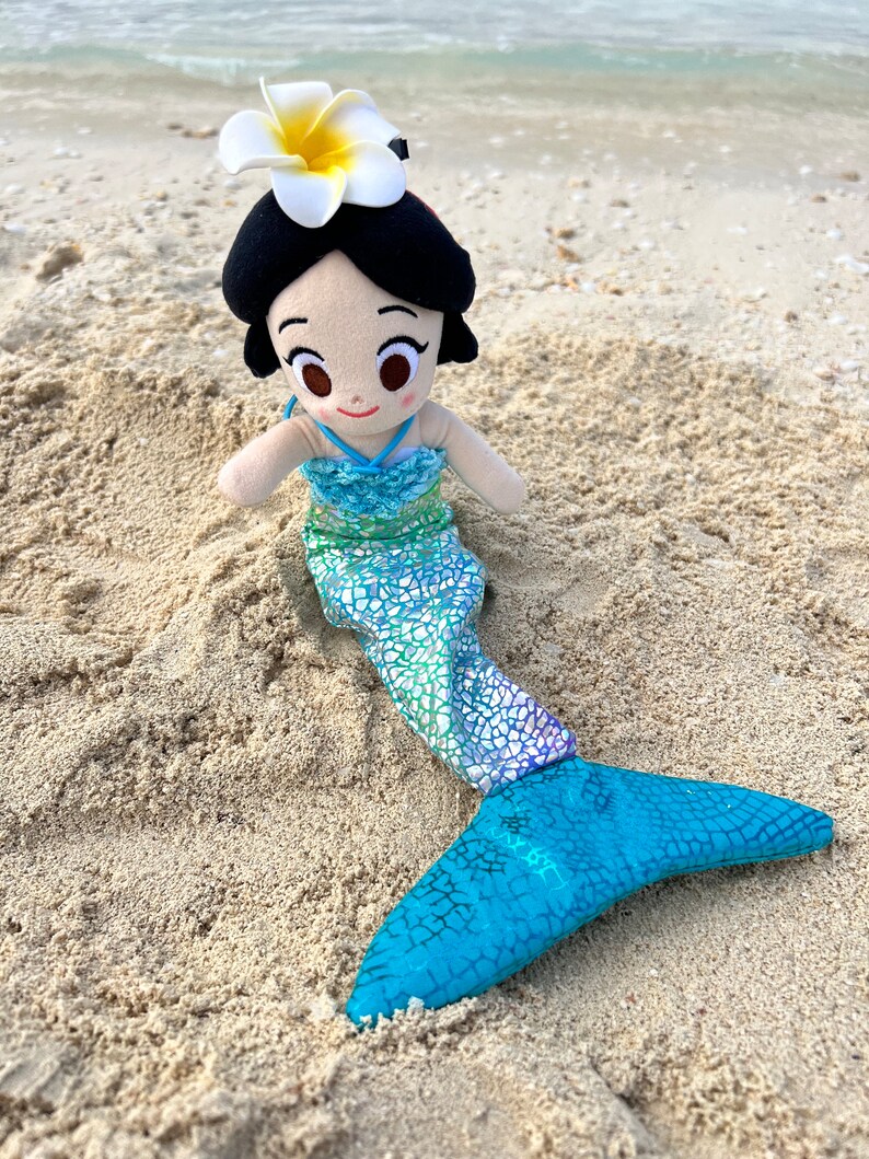 Puede incluir: Una mu&ntilde;eca de sirena de peluche con una flor blanca en la cabeza, que lleva una cola con estampado verde y azul. La mu&ntilde;eca est&aacute; sentada en la arena.