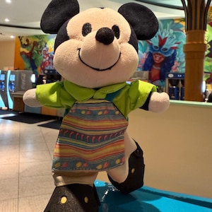 May include: A plush Mickey Mouse toy wearing a green shirt, a colorful striped apron, and black shoes. The toy is standing on a blue surface.