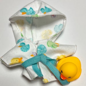K&ouml;nnte beinhalten: Ein wei&szlig;er Kapuzenbademantel mit einem blauen und gelben Entenmuster. Der Bademantel hat eine t&uuml;rkisfarbene Schleife. Eine gelbe Gummi-Ente ist im Lieferumfang enthalten.