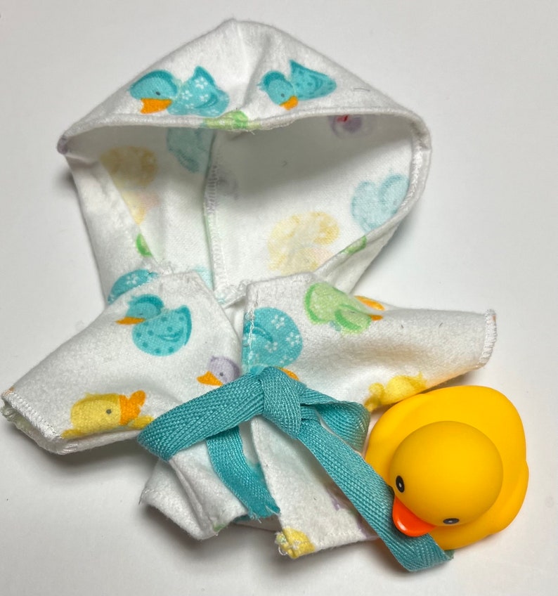 K&ouml;nnte beinhalten: Ein wei&szlig;er Kapuzenbademantel mit einem blauen und gelben Entenmuster. Der Bademantel hat eine t&uuml;rkisfarbene Schleife. Eine gelbe Gummi-Ente ist im Lieferumfang enthalten.