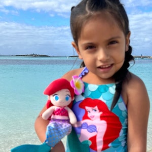 Puede incluir: Una ni&ntilde;a peque&ntilde;a lleva un traje de ba&ntilde;o azul con estampado de sirena y sostiene un juguete de peluche de sirena de pelo rojo. El traje de ba&ntilde;o tiene un patr&oacute;n de escamas verde y azul y una gran imagen de sirena en la parte delantera. La ni&ntilde;a est&aacute; de pie en una playa con agua azul de fondo.