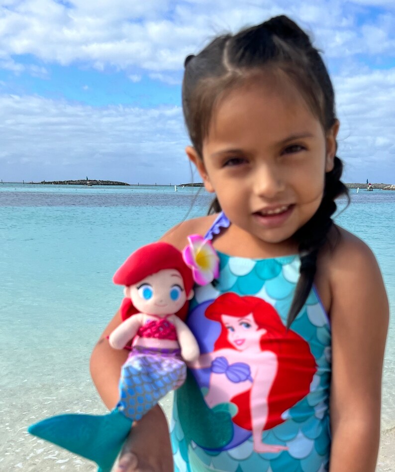 Puede incluir: Una ni&ntilde;a peque&ntilde;a lleva un traje de ba&ntilde;o azul con estampado de sirena y sostiene un juguete de peluche de sirena de pelo rojo. El traje de ba&ntilde;o tiene un patr&oacute;n de escamas verde y azul y una gran imagen de sirena en la parte delantera. La ni&ntilde;a est&aacute; de pie en una playa con agua azul de fondo.