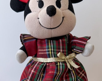 Könnte beinhalten: Eine Minnie Mouse Plüschpuppe mit einer roten Stoffschleife auf dem Kopf, bekleidet mit einem rot-grün-gelb karierten Kleid mit Rüschenärmeln und einem goldenen Schleifenbund. Die Puppe hat schwarze Ohren, ein beiges Gesicht und trägt glitzernde rote Schuhe.