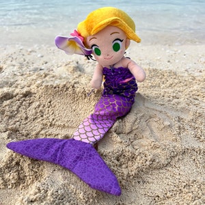 Puede incluir: Una mu&ntilde;eca de sirena de peluche con cabello largo rubio, una cola morada y una flor rosa en el cabello. La mu&ntilde;eca est&aacute; sentada en una playa de arena.