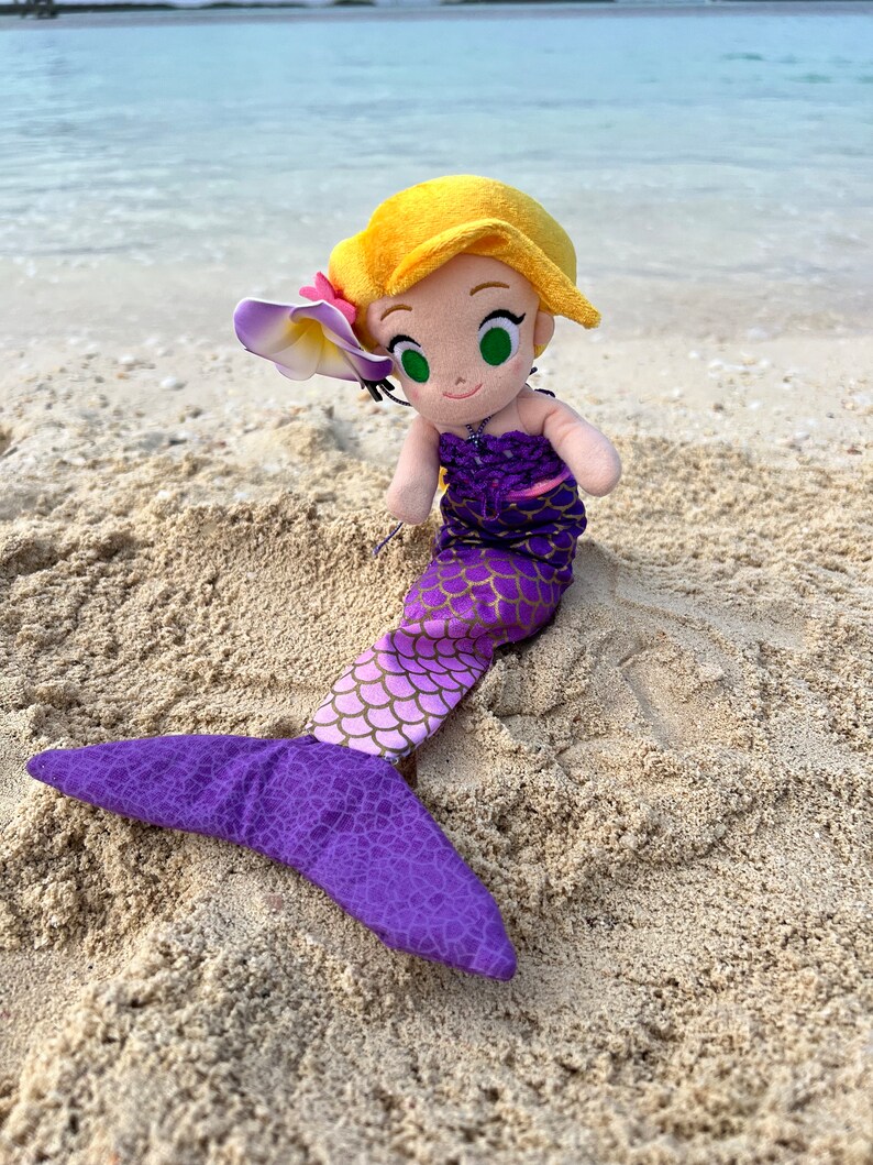 Puede incluir: Una mu&ntilde;eca de sirena de peluche con cabello largo rubio, una cola morada y una flor rosa en el cabello. La mu&ntilde;eca est&aacute; sentada en una playa de arena.