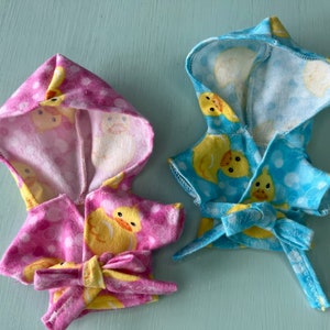 K&ouml;nnte beinhalten: Zwei Kapuzenbadem&auml;ntel f&uuml;r Puppen, einer rosa mit wei&szlig;en Punkten und einer blau mit wei&szlig;en Punkten. Beide Badem&auml;ntel haben einen gelben Gummiente-Druck.