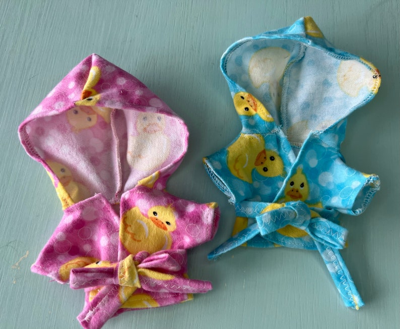 K&ouml;nnte beinhalten: Zwei Kapuzenbadem&auml;ntel f&uuml;r Puppen, einer rosa mit wei&szlig;en Punkten und einer blau mit wei&szlig;en Punkten. Beide Badem&auml;ntel haben einen gelben Gummiente-Druck.