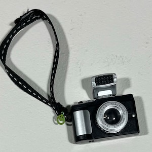 May include: A black and silver toy camera with a black strap and a green smiley face charm. The camera has a flash and a lens that says "ZHAOXIANGLI OPTICAL 1:3.5/6 14-24mm".