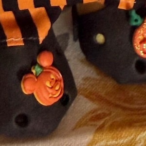 May include: A pair of black clogs featuring orange and black striped fabric cuffs. Each clog is decorated with a small, orange, pumpkin-shaped charm with a green stem and mouse-ear silhouette, accented with gold glitter on the surface.