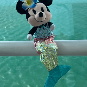 Puede incluir: Un juguete de peluche de Mickey Mouse vestido como una sirena. El juguete tiene una cabeza blanca y negra con orejas grandes, una flor rosa y blanca en la cabeza y una cola de sirena azul y verde.