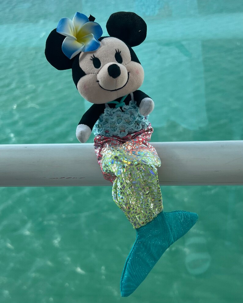 Puede incluir: Un juguete de peluche de Mickey Mouse vestido como una sirena. El juguete tiene una cabeza blanca y negra con orejas grandes, una flor rosa y blanca en la cabeza y una cola de sirena azul y verde.