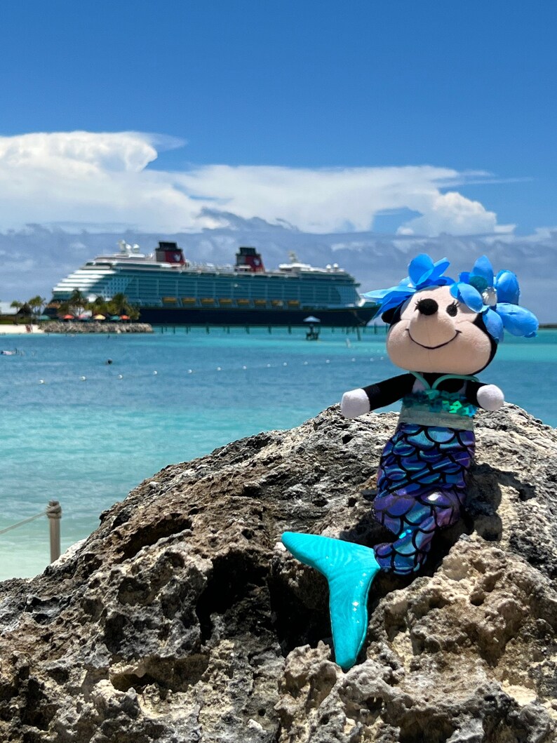 Puede incluir: Un juguete de peluche de sirena azul y morado con una corona de flores azules se sienta en una roca con un gran crucero en el fondo.