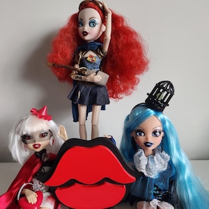 Könnte beinhalten: Drei Modepuppen mit unterschiedlichen Frisuren und Outfits. Die Puppe links hat weißes Haar, einen roten Umhang und ein weißes Kleid. Die Puppe in der Mitte hat rotes Haar, ein blaues und schwarzes Kleid und eine braune Tasche. Die Puppe rechts hat blaues Haar, ein blaues und weißes Kleid und einen schwarzen Vogelkäfig-Hut.