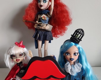 May include: Three fashion dolls with different hairstyles and outfits. The doll on the left has white hair, a red cape, and a white dress. The doll in the middle has red hair, a blue and black dress, and a brown bag. The doll on the right has blue hair, a blue and white dress, and a black birdcage hat.