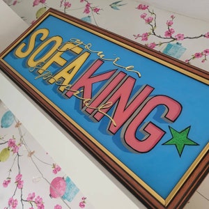 May include: A framed wall art piece with the phrase "You're the Sofa King" in a vibrant, cartoon style. The letters are yellow and pink with grey outlines, set against a blue backdrop, framed in brown and gold. A green star is in the corner.