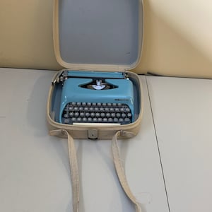 Può includere: Una macchina da scrivere vintage azzurra con tasti neri è all'interno di una custodia aperta color bianco sporco. La custodia ha una tracolla abbinata color bianco sporco. È un classico articolo per l'ufficio.