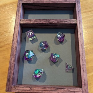 Puede incluir: Una bandeja de dados de madera con un interior de fieltro gris. La bandeja contiene siete dados iridiscentes con n&uacute;meros blancos. Los dados son un d4, d6, d8, d10, d12 y d20.