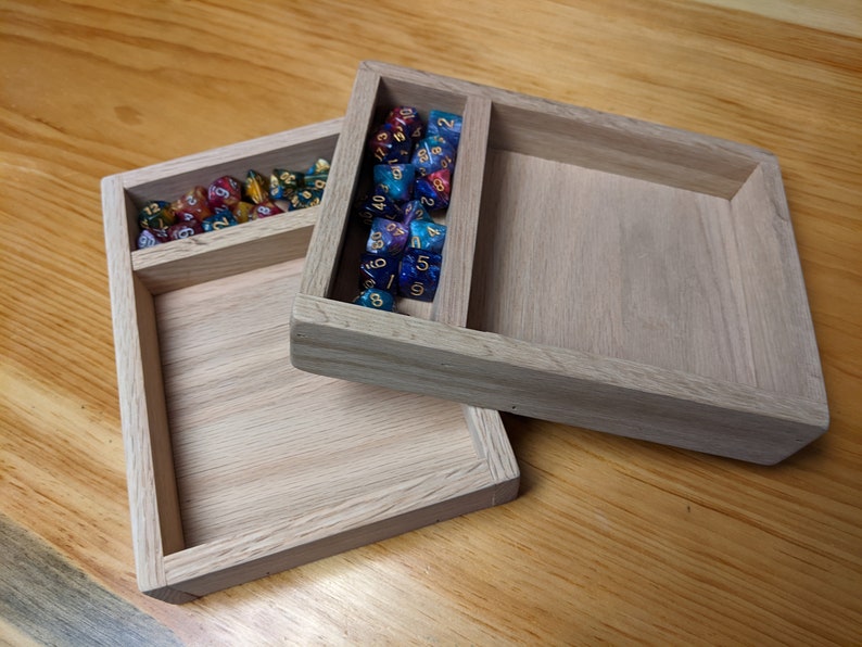 Puede incluir: Dos bandejas de madera con un compartimento para dados. La bandeja superior tiene una variedad de dados coloridos con n&uacute;meros dorados. La bandeja inferior est&aacute; vac&iacute;a.