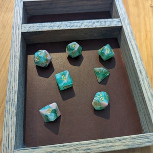 Puede incluir: Un conjunto de siete dados verdes y rosas con n&uacute;meros dorados en una bandeja de madera. Los dados son de varias formas, incluyendo un d4, d6, d8, d10, d12 y d20.