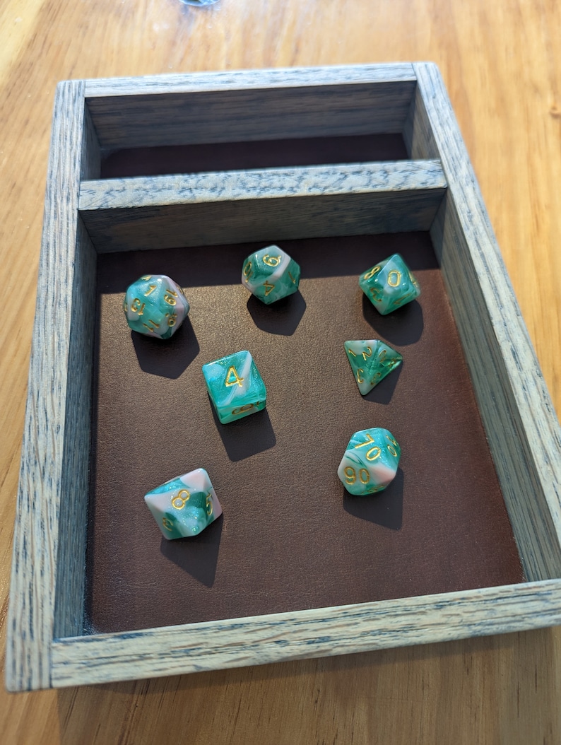 Puede incluir: Un conjunto de siete dados verdes y rosas con n&uacute;meros dorados en una bandeja de madera. Los dados son de varias formas, incluyendo un d4, d6, d8, d10, d12 y d20.