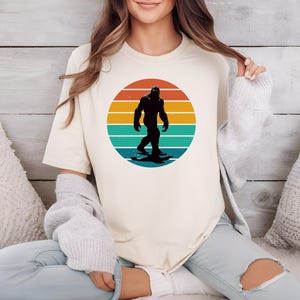 May include: A cream-colored t-shirt with a retro-style graphic of a Bigfoot walking across a sunset. The graphic is in shades of orange, yellow, blue, and green.