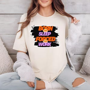 Könnte beinhalten: Ein weißes T-Shirt mit einer schwarzen und orangenen Grafik, auf der "Born to Sleep Forced to Work" steht.