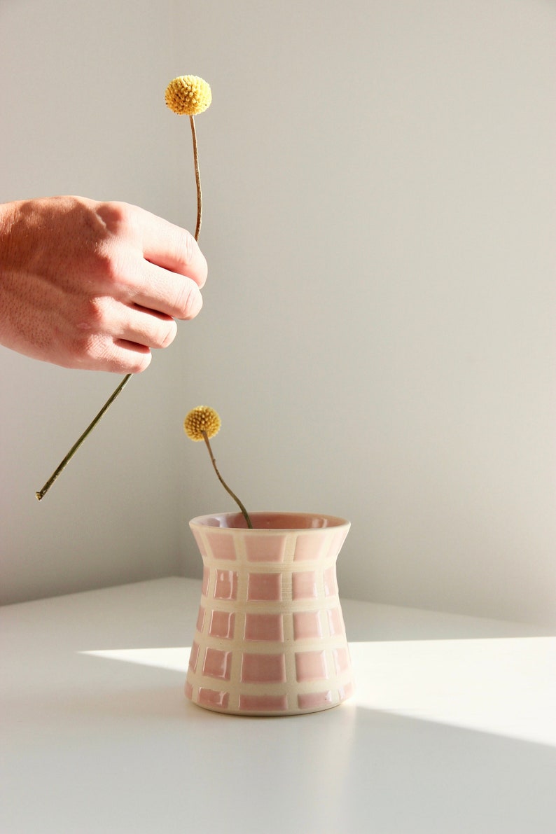 A hand putting a yellow billy ball flower into a retro-modern vase, with pink squares.