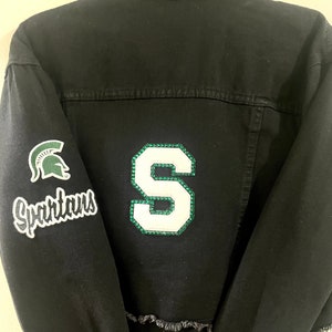Puede incluir: Chaqueta vaquera negra con un parche 'S' blanco y verde en la espalda y un parche verde con un casco espartano y 'Spartans' en la manga izquierda.