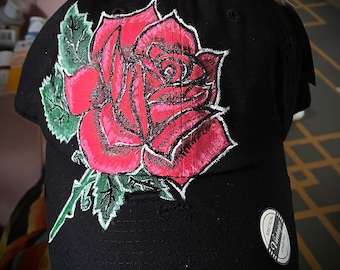 May include: A black baseball cap with a hand-painted red rose design. The rose has green leaves and is centered on the front of the cap. The cap has a distressed look with a small tear on the front.
