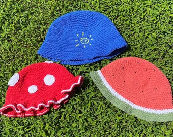 May include: Three handmade crochet bucket hats on green grass. One is blue with a yellow embroidered sun, another is red with white spots, and the third is watermelon-themed with red, green, and white colours.