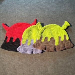 May include: Three crocheted hats in different colors. The first hat is red with black dripping down the sides. The second hat is green with purple dripping down the sides. The third hat is green with brown dripping down the sides.