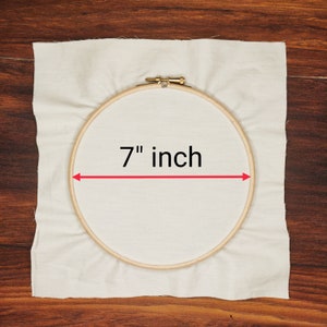 K&ouml;nnte beinhalten: Ein h&ouml;lzerner Stickrahmen mit einem Durchmesser von etwa 17,78 cm liegt auf einem cremefarbenen Stoff. Der Text "7 inch" ist zu sehen, mit einem roten Pfeil, der das Ma&szlig; anzeigt.