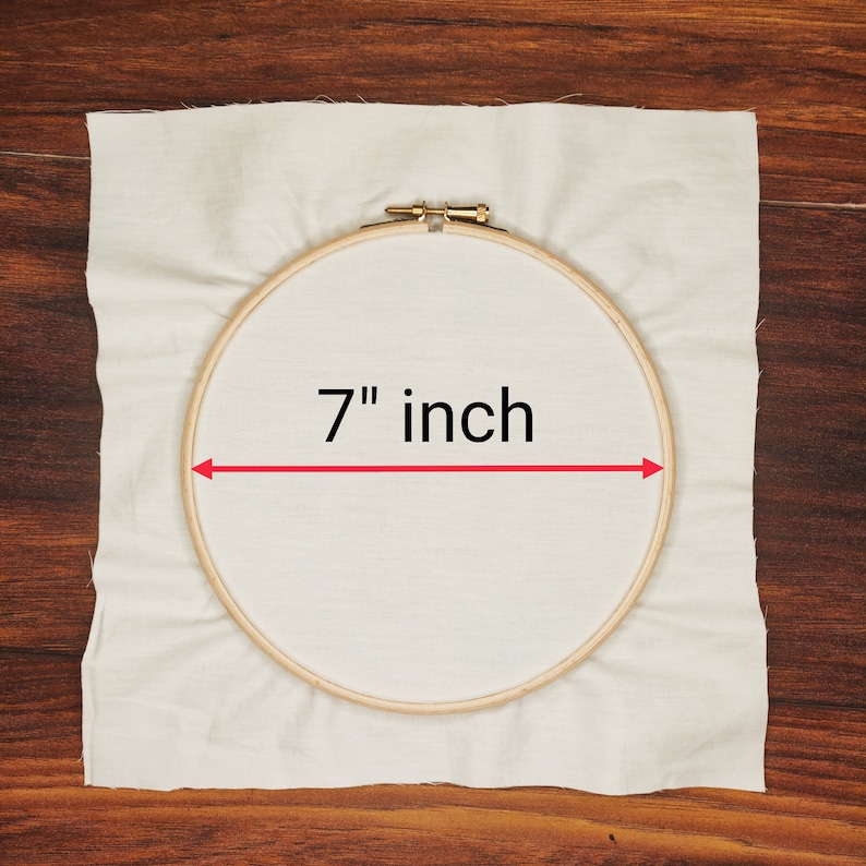 K&ouml;nnte beinhalten: Ein h&ouml;lzerner Stickrahmen mit einem Durchmesser von etwa 17,78 cm liegt auf einem cremefarbenen Stoff. Der Text "7 inch" ist zu sehen, mit einem roten Pfeil, der das Ma&szlig; anzeigt.