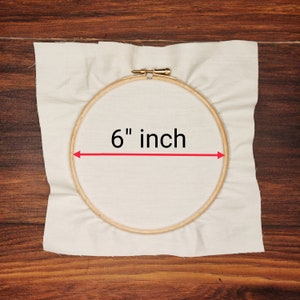 K&ouml;nnte beinhalten: Ein h&ouml;lzerner Stickrahmen mit einem Durchmesser von 15,24 cm, der ein St&uuml;ck cremefarbenen Stoff h&auml;lt. Der Text "6 inch" ist in Schwarz dargestellt, mit einem roten Pfeil, der das Ma&szlig; angibt. Der Hintergrund ist eine Holzoberfl&auml;che.