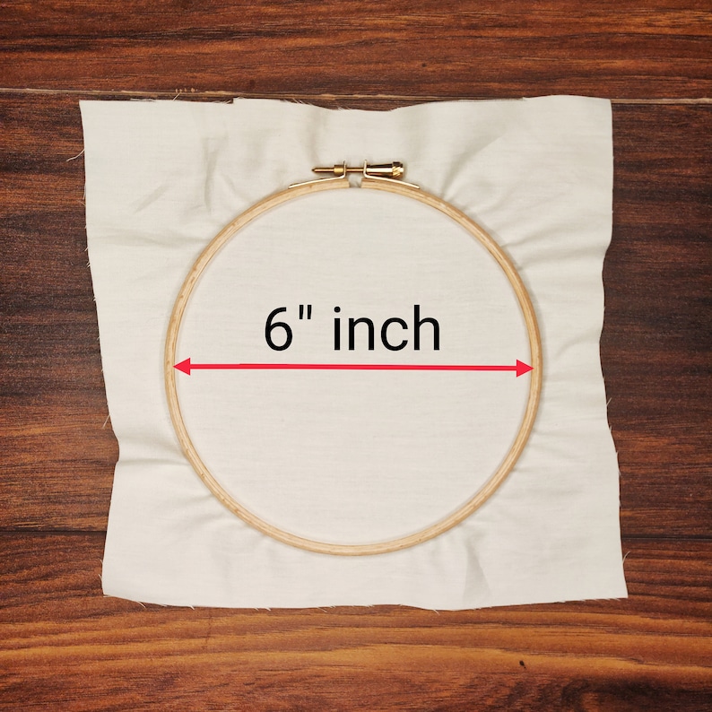 K&ouml;nnte beinhalten: Ein h&ouml;lzerner Stickrahmen mit einem Durchmesser von 15,24 cm, der ein St&uuml;ck cremefarbenen Stoff h&auml;lt. Der Text "6 inch" ist in Schwarz dargestellt, mit einem roten Pfeil, der das Ma&szlig; angibt. Der Hintergrund ist eine Holzoberfl&auml;che.
