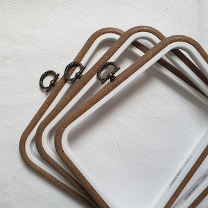 May include: Four white and brown rectangular trays with decorative handles. The trays have a white interior and a brown, wood-grain-like border. The handles are a dark bronze colour. The trays are stacked on top of each other.