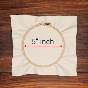 K&ouml;nnte beinhalten: Ein h&ouml;lzerner Stickrahmen mit einem Durchmesser von 12,7 cm ist auf einem St&uuml;ck cremefarbenem Stoff zentriert. Der Stoff ist leicht zerknittert und liegt auf einer Holzoberfl&auml;che. Der Text "5 inch" wird in Schwarz angezeigt.