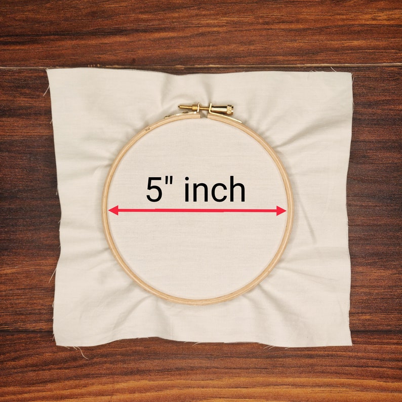 K&ouml;nnte beinhalten: Ein h&ouml;lzerner Stickrahmen mit einem Durchmesser von 12,7 cm ist auf einem St&uuml;ck cremefarbenem Stoff zentriert. Der Stoff ist leicht zerknittert und liegt auf einer Holzoberfl&auml;che. Der Text "5 inch" wird in Schwarz angezeigt.