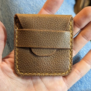 May include: A small, brown leather wallet with a textured surface. The wallet has a flap closure and a card slot on the front. The edges are stitched with yellow thread. The wallet is designed to hold cards and cash.