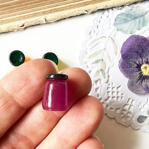 May include: A miniature, translucent, magenta-colored jar with a dark green lid. Two dark green and one orange circular objects are visible. A pressed purple pansy flower and a decorative paper doily are in the background.