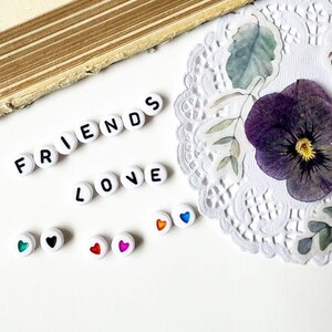 Könnte beinhalten: Weiße Perlen schreiben "FRIENDS" und "LOVE" mit schwarzen Buchstaben. Kleine weiße Perlen mit bunten Herzmotiven sind ebenfalls vorhanden. Ein getrocknetes violettes Stiefmütterchen und eine dekorative Serviette sind im Hintergrund.
