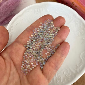 Op de afbeelding: Een handvol iriserende glazen kralen in verschillende pasteltinten van roze, blauw en geel. De kralen zijn klein en rond en hebben een subtiele glans.