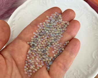 Könnte beinhalten: Eine Handvoll irisierende Glasperlen in verschiedenen Pastelltönen von Rosa, Blau und Gelb. Die Perlen sind klein und rund und haben einen dezenten Schimmer.