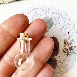 May include: A miniature clear glass bottle with a cork stopper. The bottle has a bulbous base and a slender neck. The bottle is held in a hand, with a white lace doily and floral stickers in the background.