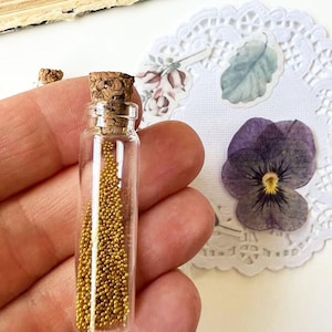 May include: A small, clear glass bottle with a cork stopper, filled with tiny gold beads. The bottle is held in a hand, with a dried purple flower and decorative paper doily in the background.