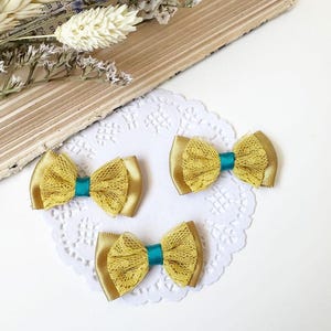 Peut inclure: Trois nœuds pour cheveux dorés avec des centres en ruban turquoise et un revêtement en dentelle. Les nœuds sont disposés sur une nappe en dentelle blanche, avec un livre vintage et des fleurs séchées en arrière-plan. Accessoire de cheveux décoratif.