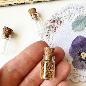 May include: Miniature glass bottles with cork stoppers, one filled with tiny gold beads. The bottles are displayed on a white surface with floral embellishments and a purple flower. The bottles are small, likely for decorative purposes.