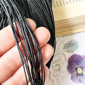 May include: Close-up of a hand holding a bundle of black waxed cord. The cord appears to be tightly woven and has a slightly shiny finish. The background includes a vintage book page and floral decorations.