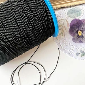 May include: A spool of black thread with a blue rim. The thread is tightly wound, with a strand extending from the spool. Decorative elements, including a purple flower and lace, are in the background.