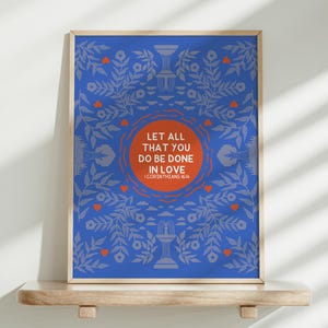 May include: A framed art print with the text "LET ALL THAT YOU DO BE DONE IN LOVE" in an orange circle. The blue background has a floral pattern with orange hearts and grey accents. The print is displayed on a wooden shelf.